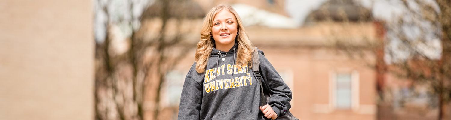A female student in a gray Kent State sweatshirt walks with a backpack in front of a building