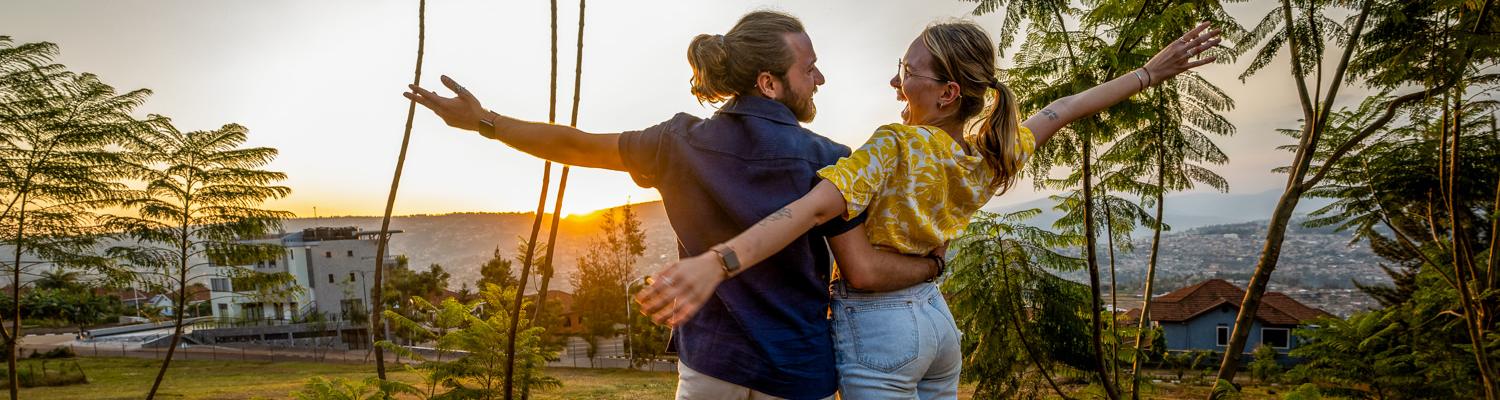 A golden-hour medium shot of a man and a woman standing in a grassy area with their backs partially to the camera, embracing and looking at each other joyfully with arms wide open. The setting sun glows behind them through thin, tall trees, overlooking a valley with houses scattered across the hills in the background.
