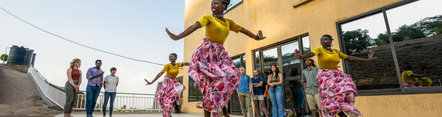 A wide shot of three Rwandan women performing a traditional dance on a polished, reflective outdoor patio next to a modern yellow building. They are wearing bright yellow tops and vibrant pink-and-white floral skirts, captured mid-movement with arms outstretched. In the background, a group of students and faculty members stand watching the performance.