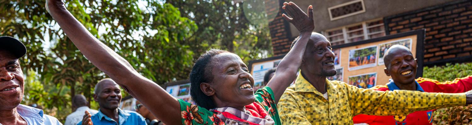 A vibrant, close-up shot capturing a moment of celebration. A Rwandan woman in a green floral dress has both arms raised high in the air, smiling brightly. Surrounding her are several other men and women laughing and cheering, with a wooden display board featuring photographs visible in the background.