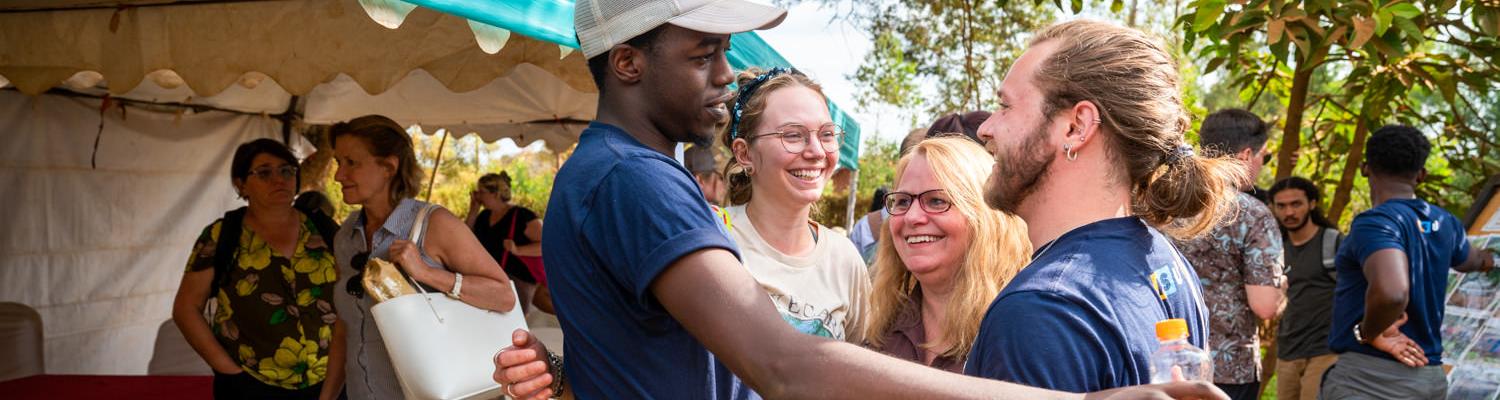 A joyful group interaction outdoors under a bright blue-and-white canopy. In the foreground, a young Rwandan man wearing a baseball cap and a dark blue shirt is embracing or greeting a Caucasian man with long hair tied back, who is also wearing a �Ը������� University blue shirt. Surrounding them are other smiling students and faculty members.