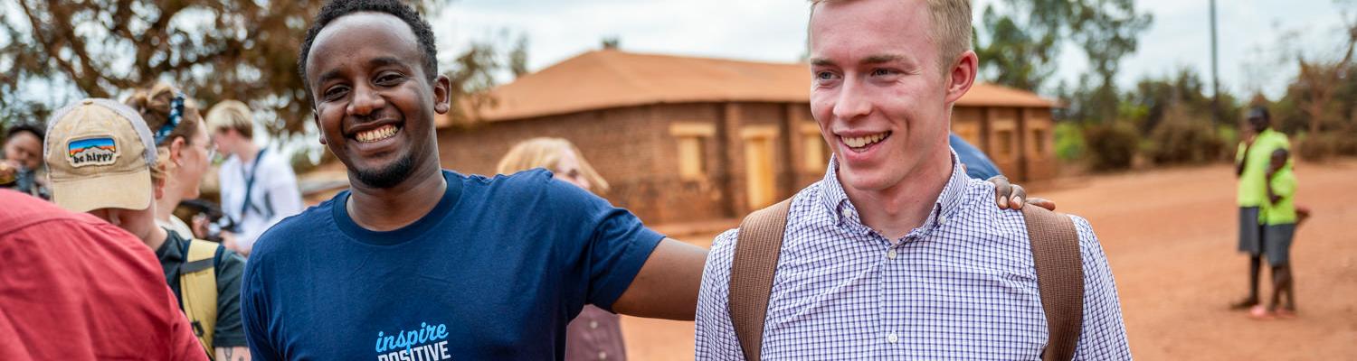 candid medium shot of two young men smiling and walking together on a dirt road. On the left, a Rwandan man in a dark blue T-shirt that reads "inspire POSITIVE CHANGE" smiles broadly with his arm around a blond Caucasian man wearing a purple gingham button-down shirt and a backpack. Other people and a simple brick building are visible in the background.