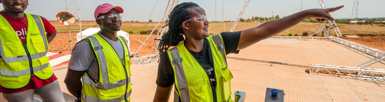 A medium shot of three individuals standing on an outdoor launch pad under a clear sky, surrounded by metal lattice towers. A Rwandan woman in the center, wearing safety glasses and a neon yellow vest, smiles as she points into the distance. Two men in safety vests stand behind her, tracking where she is pointing.