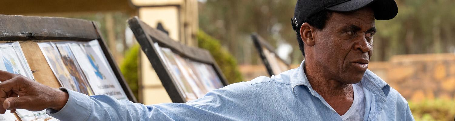 A man in a black cap and light blue shirt gestures toward a display of framed prints and artwork at an outdoor art market in Rwanda.