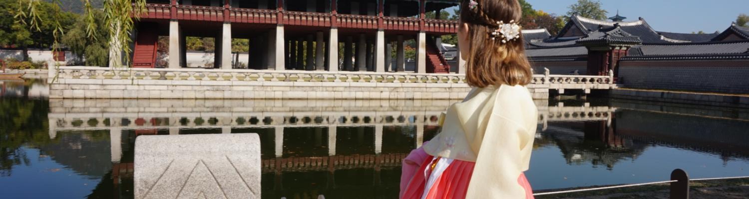 A student dressed in hanbok at a palace in South Korea. There is a historical building surrounded by water.
