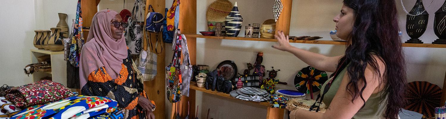 A visitor browses handmade goods with a shopkeeper inside a Rwandan artisan shop, with shelves stocked with woven baskets, ceramics, textiles, and local artwork filling the background.