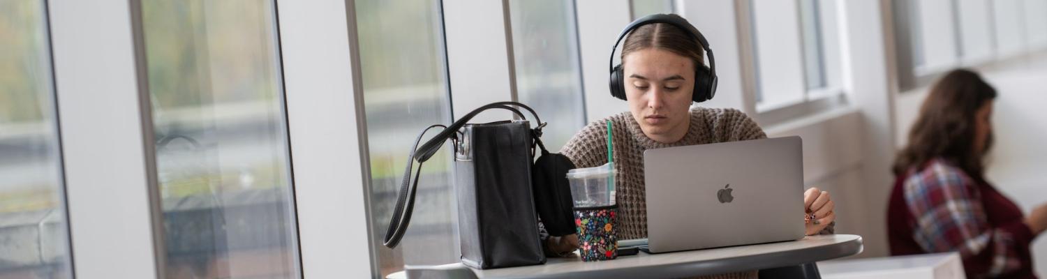 Female student sitting at a table and studying on her laptop, with a wall of windows behind her