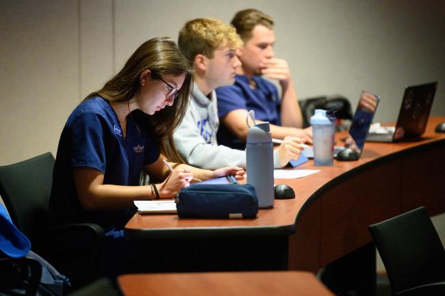 A group of three students taking notes during a lecture indoors