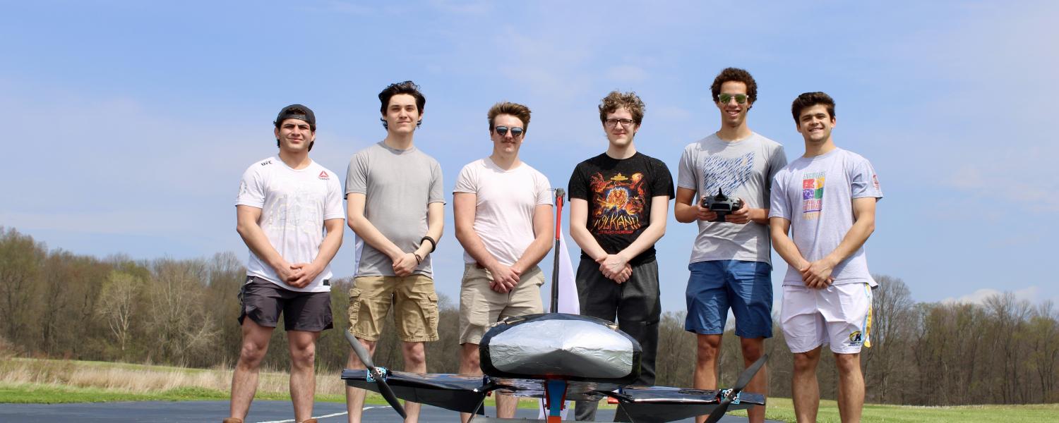 2026 Design, Build, Fly team consisting of six male college students standing on the runway behind their finished aircraft