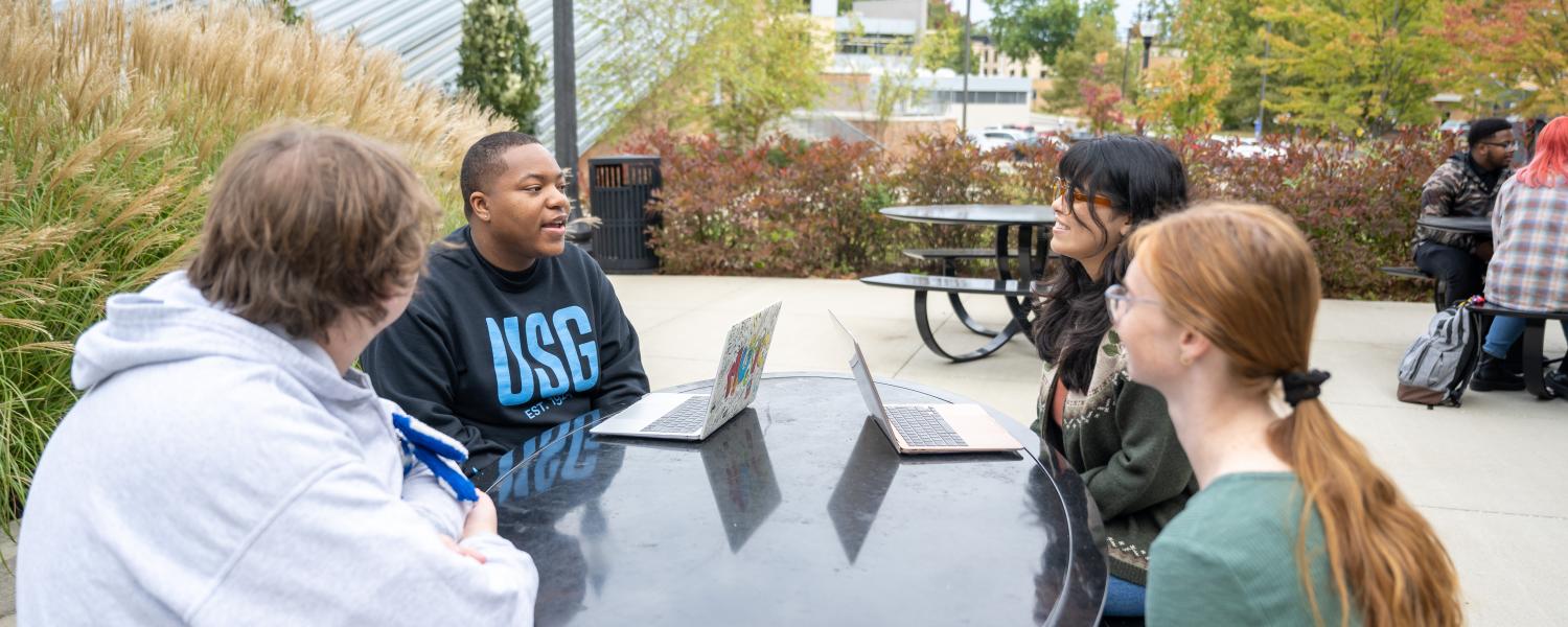 Four students sit around a table outside on campus discussing their work.