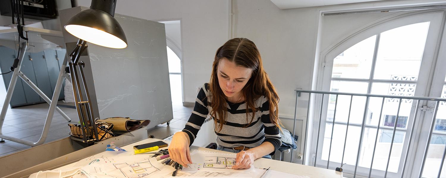 A photo of a young woman working at a desk strewn with papers