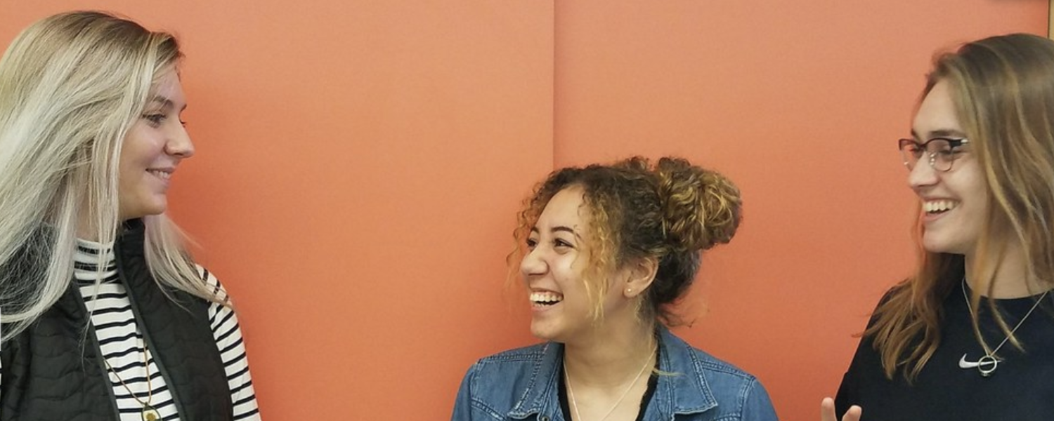 Three American Sign Language/English Interpreting students stand against a colorful wall. The girl on the far right is signing with both hands.