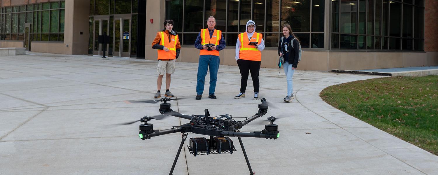 Four people standing at a distance wearing high-visibility vests with a medium-sized quadcopter in the foreground.