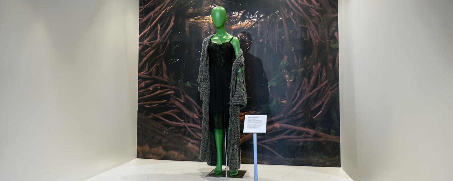Long gray sweater on a green mannequin positioned in front of a backdrop with vines. 