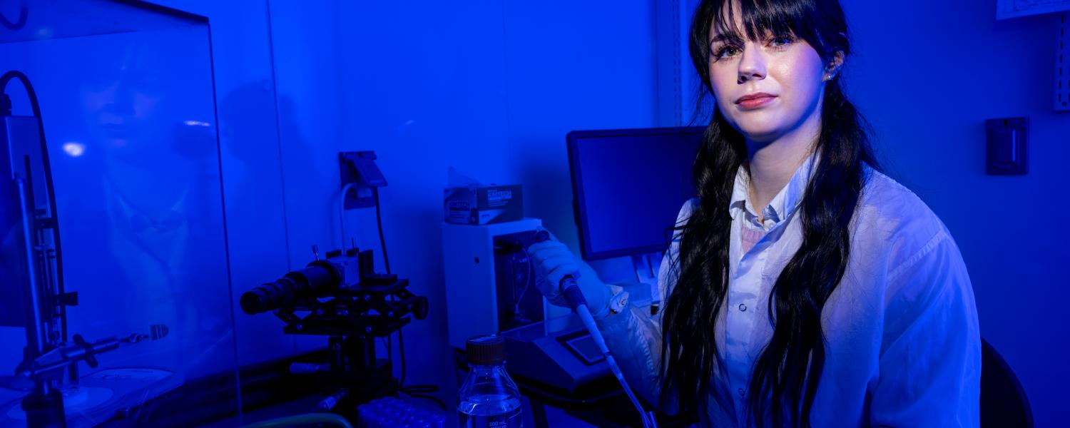 A student wearing a white lab coat stands in a dark room, surrounded by beakers and other scientific equipment.