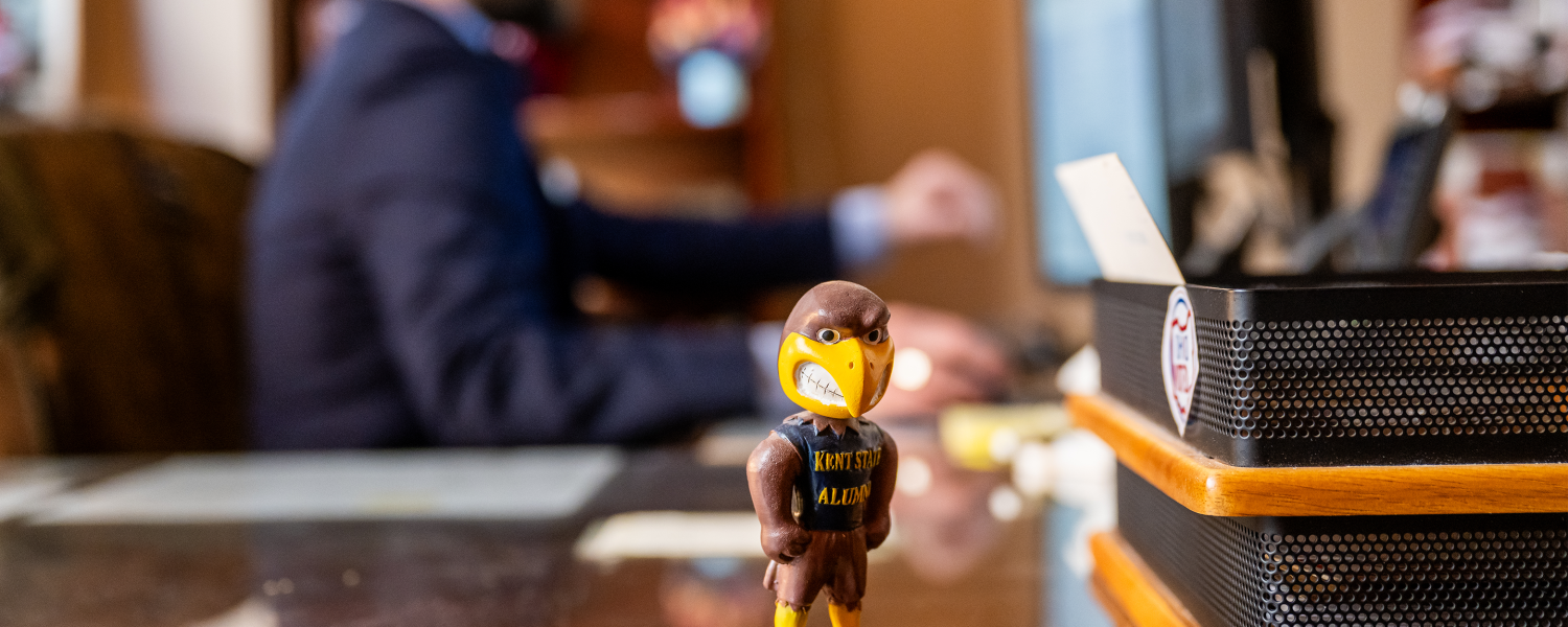 A bobblehead of ����Թ� State's mascot Flash the Golden Eagle sits on a desk with someone working on a computer out of fucus in the background