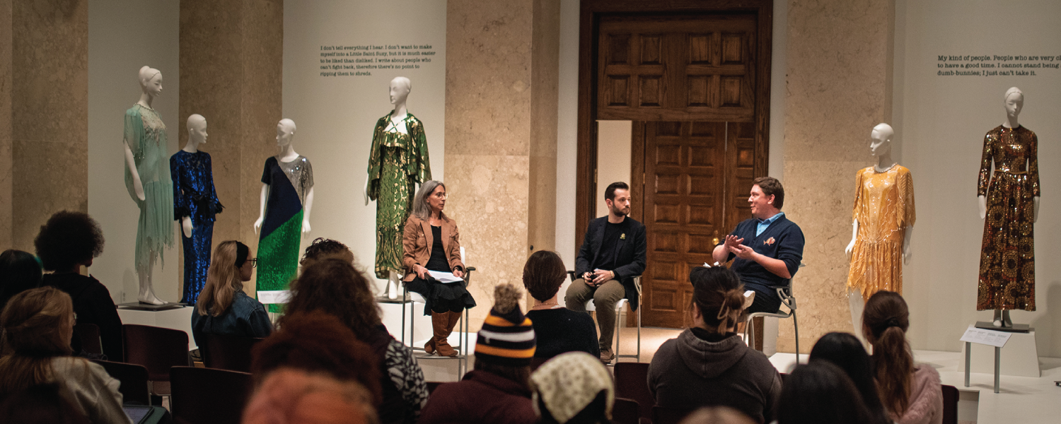 Three people discuss on center stage with clothed mannequins dressed in sequined garments standing around them as the speakers sit in front of an audience