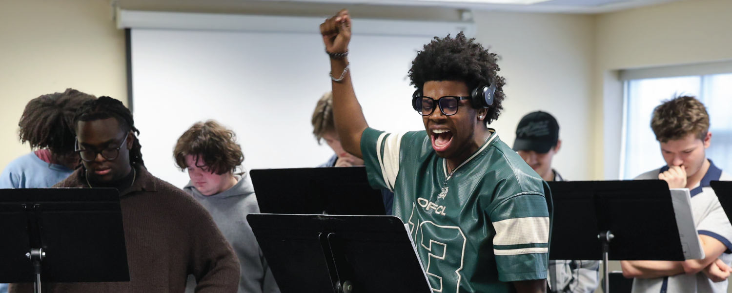 Student with headphones raises his hand passionately as he sings a note from the sheet music displayed in front of him