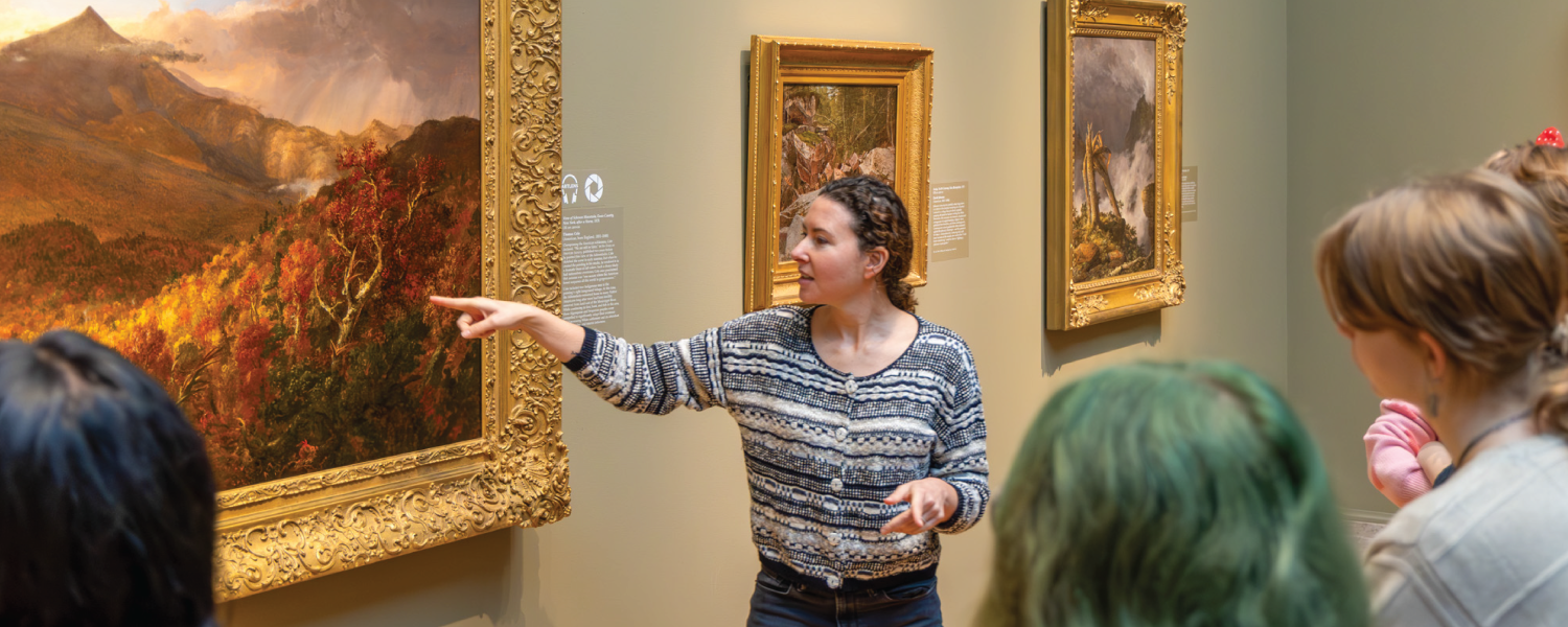 Shana points to a landscape painting with a golden frame before a group of students