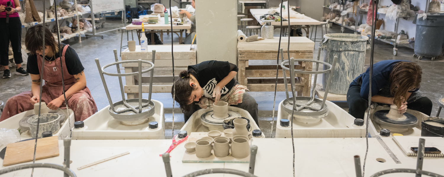 Three ceramic artists perform pottery wheel techniques side-by-side across a long table