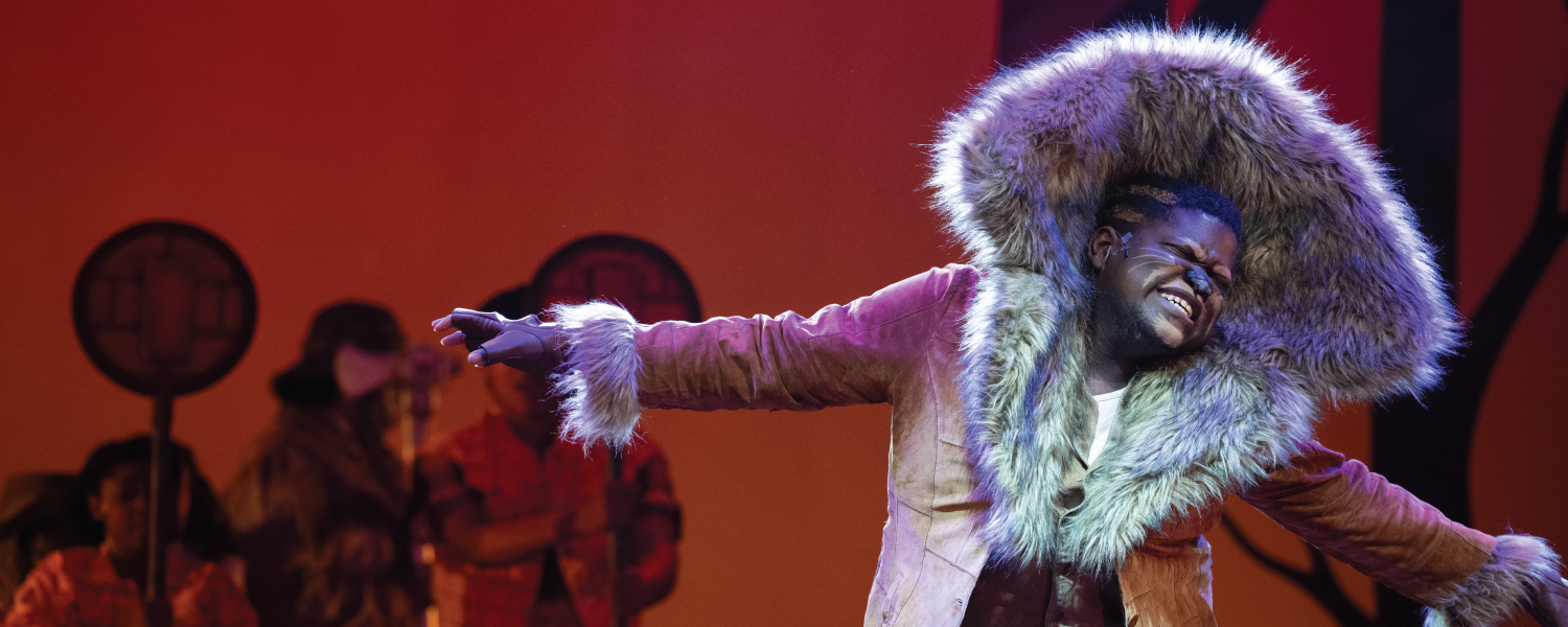 Actor playing part of the Lion sings and spreads his arms, a large fur fabric splaying upwards from his shoulders to around his head