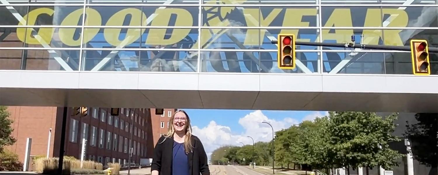 A woman standing on a sunny street with an elevated bridge behind her reading "Goodyear"