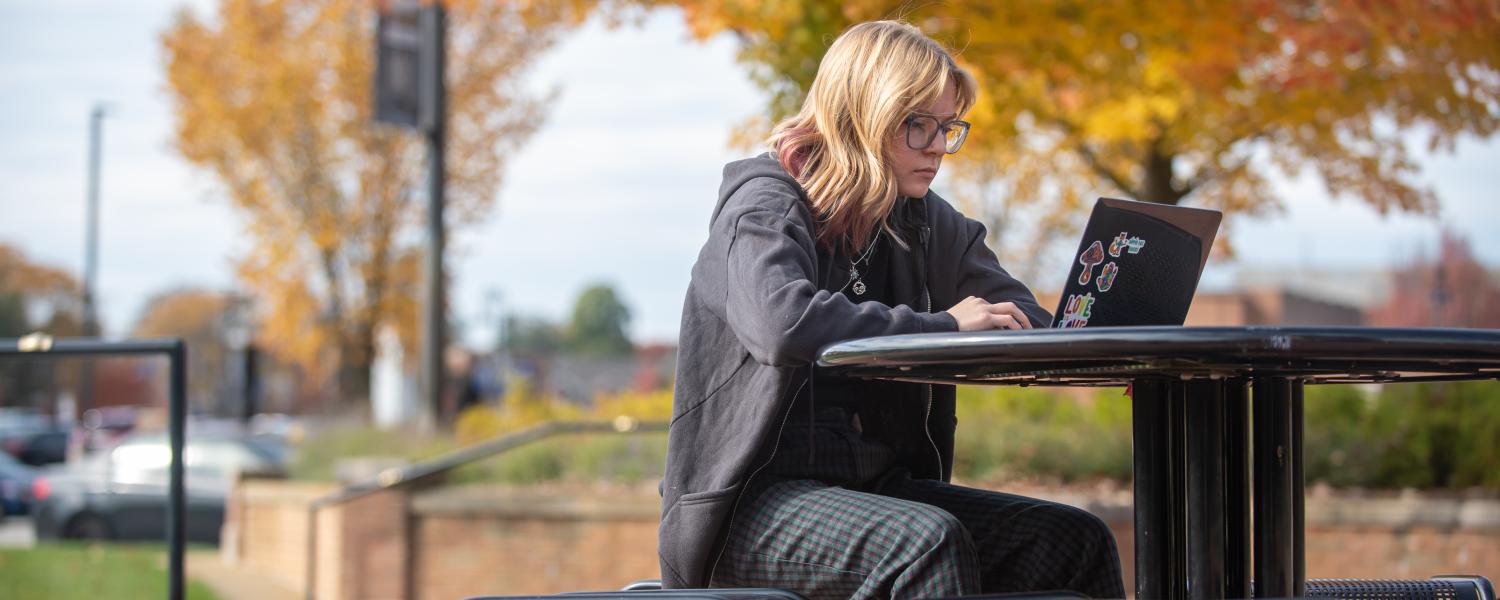 A student sits at a picnic table outdoors, working on a laptop.