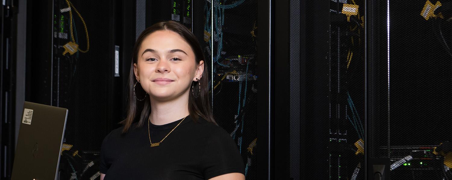 A student smiling proudly while holding a laptop, standing in front of physical database equipment.