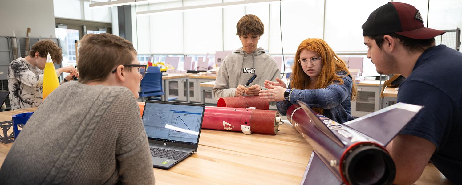 several students seated around a work table with a partially disassembled model rocket