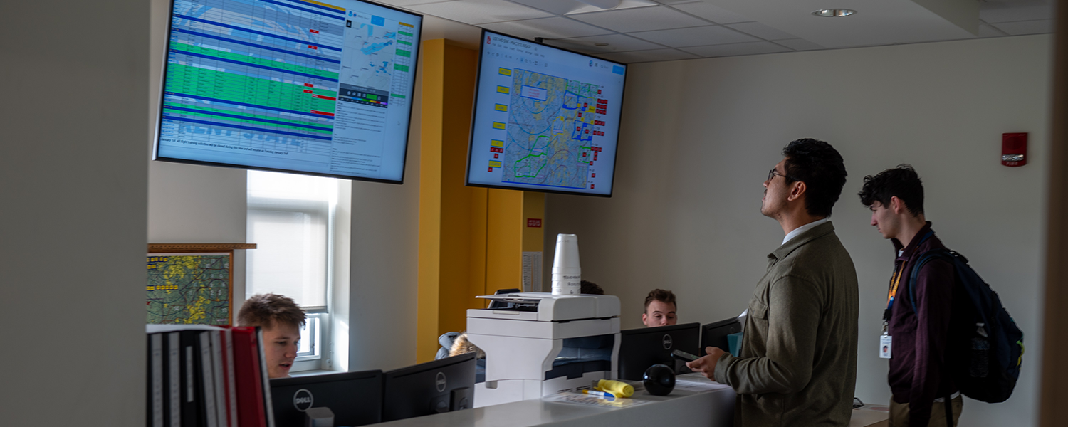 Two students across a counter looking at two large screens showing arrival and departure times for student flights.