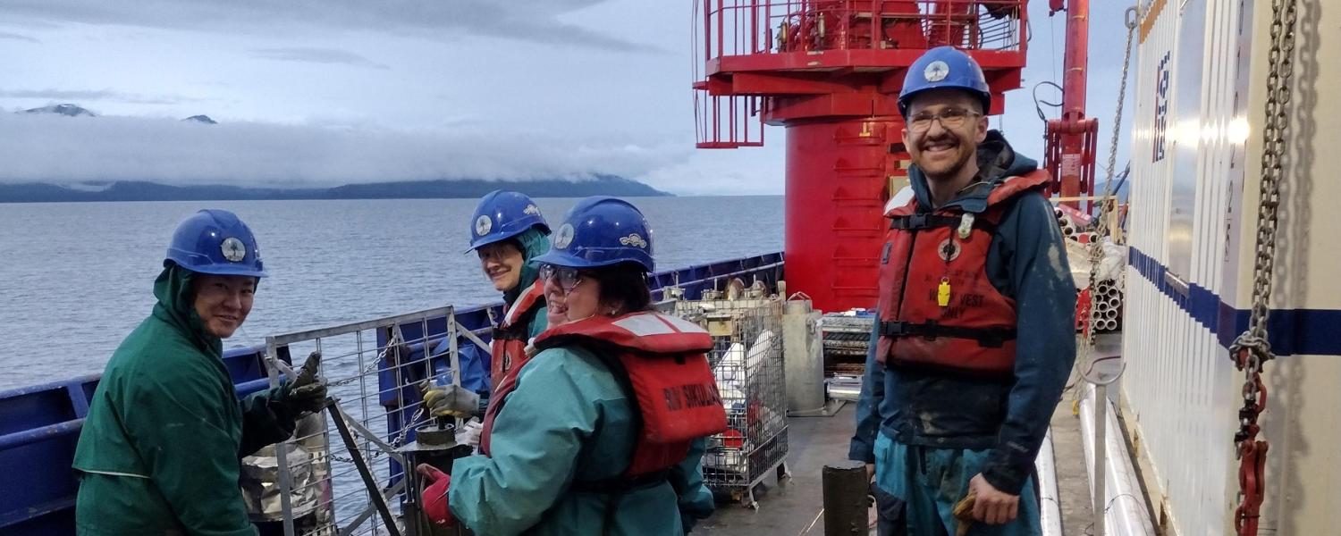 Four people smiling and standing on a boat wearing rain gear and life preservers, next to some equipment, with water and a gray sky in the background.
