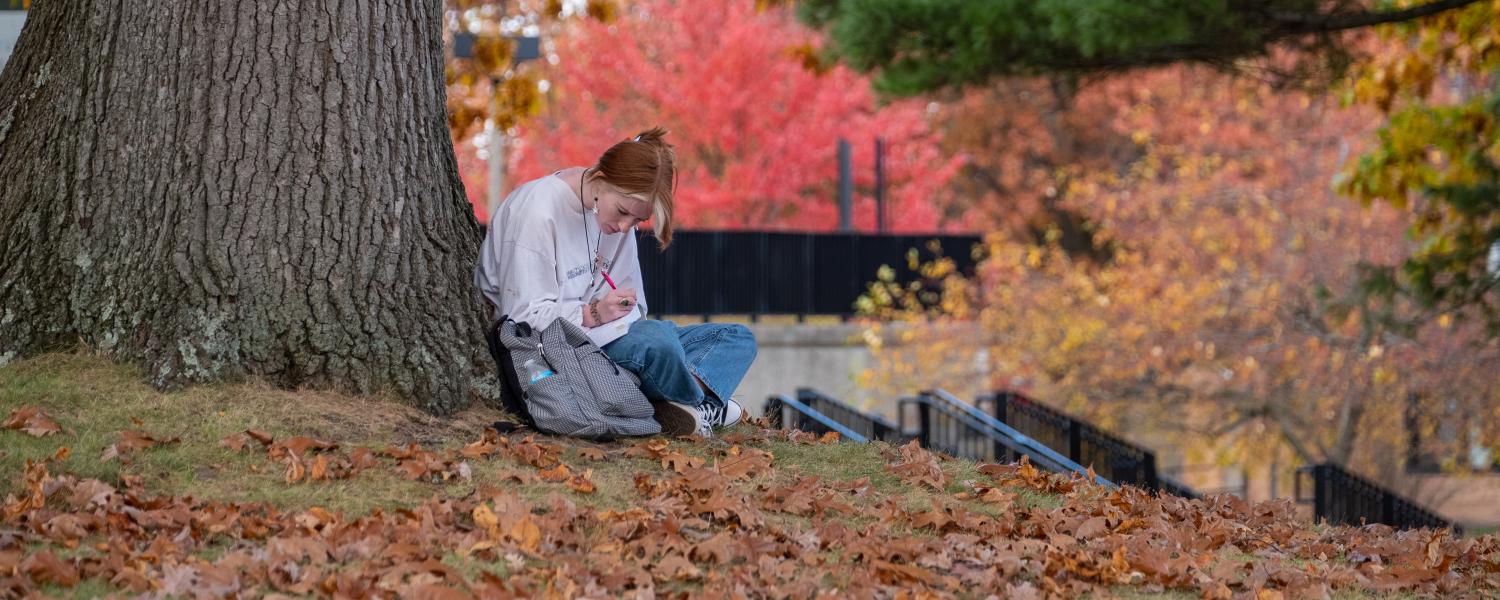 A student sits in front of a tree in the fall, writing with a pen and paper.