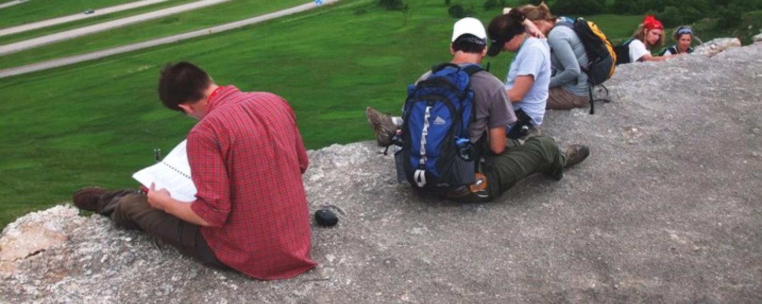 Students sit atop a high rock, reviewing notes with backpacks on, overlooking a green landscape and winding roads.