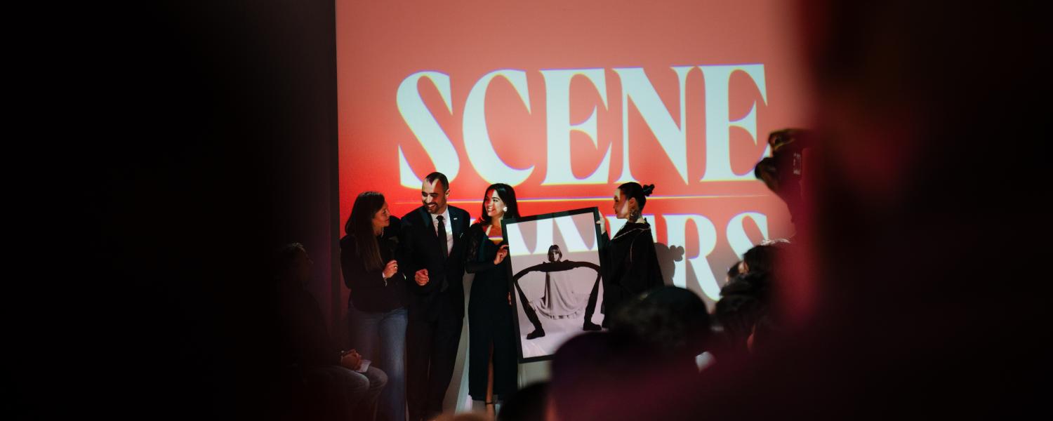 Silhouettes of New York City fashion show participants stand in front of a red digital screen before an audience