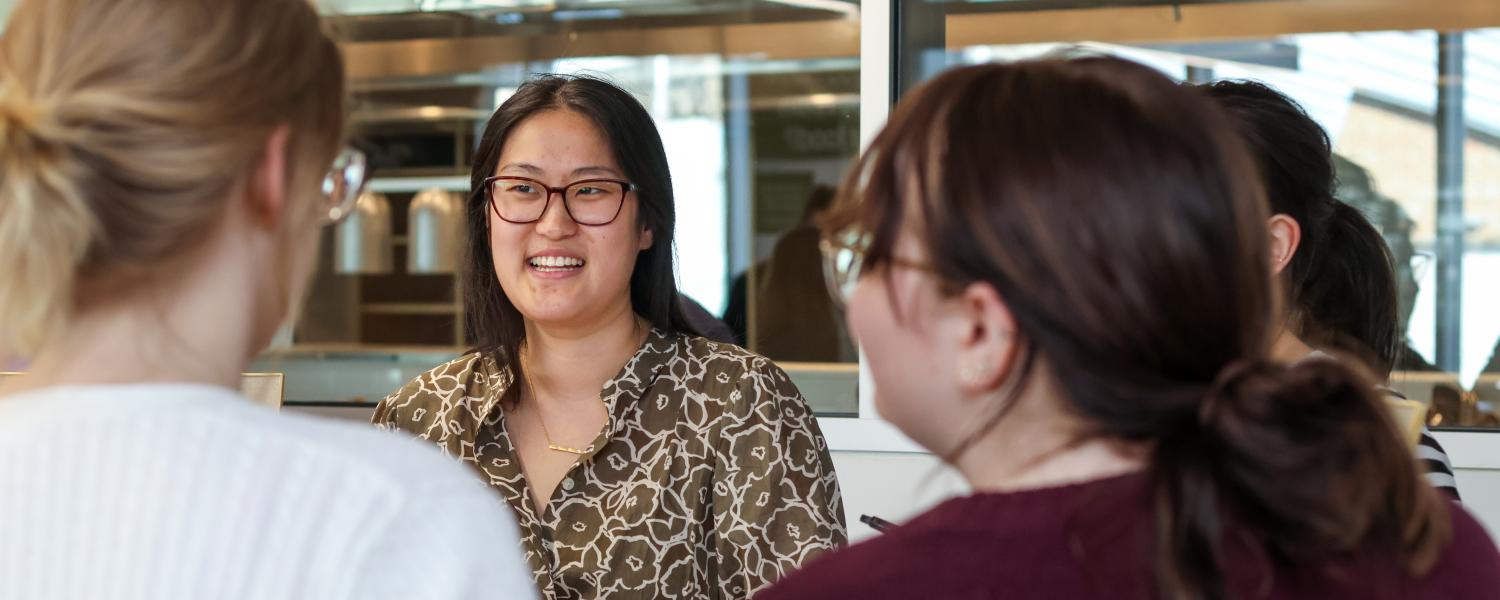A woman in glasses and a patterned blouse smiles while speaking with two people standing across from her at a career fair.