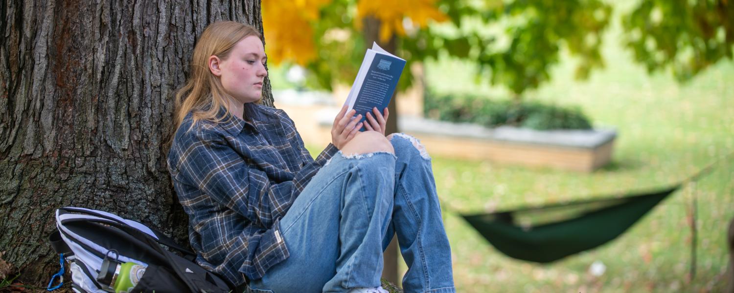 A student sits with their back against a tree, reading a book beside their backpack, while another person relaxes in a hammock in the distance.
