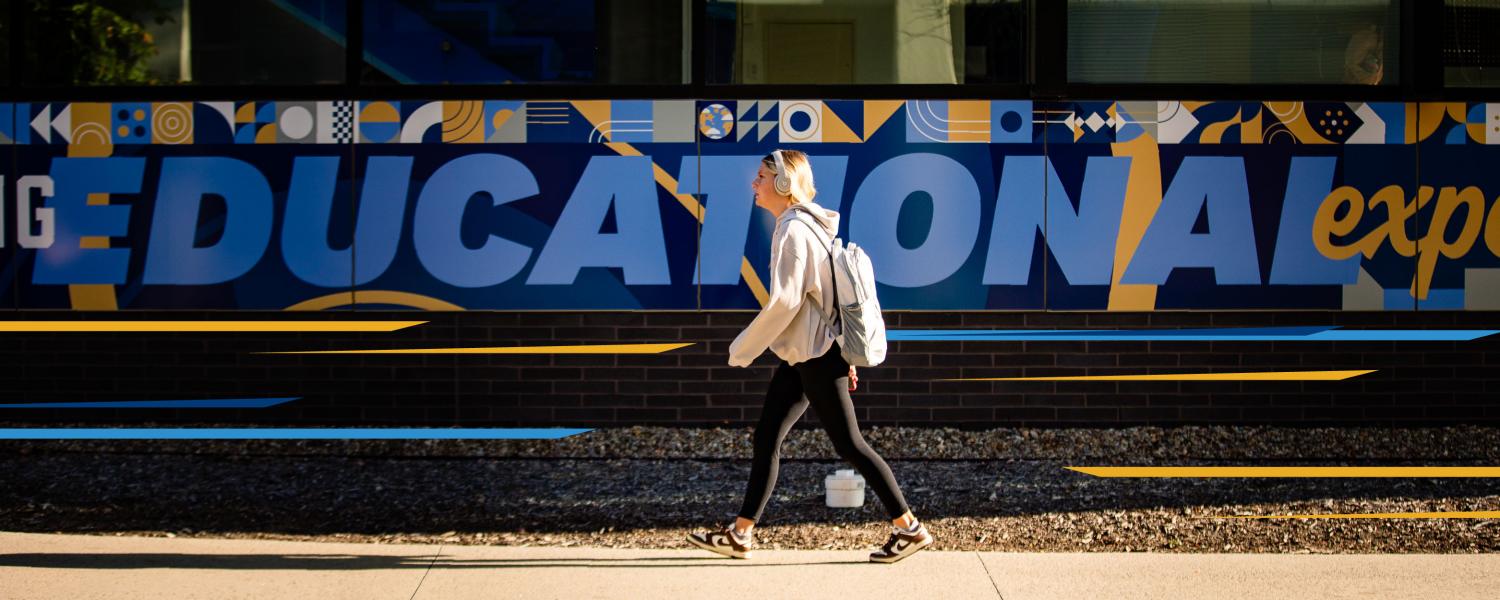 A student walks front left to right in front of a sign that reads "education" on campus.