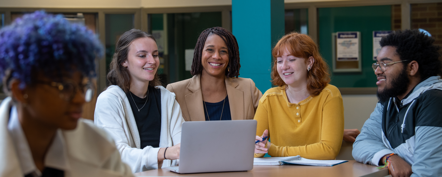 A small group of students sit around a table as they smile and work on a laptop together.