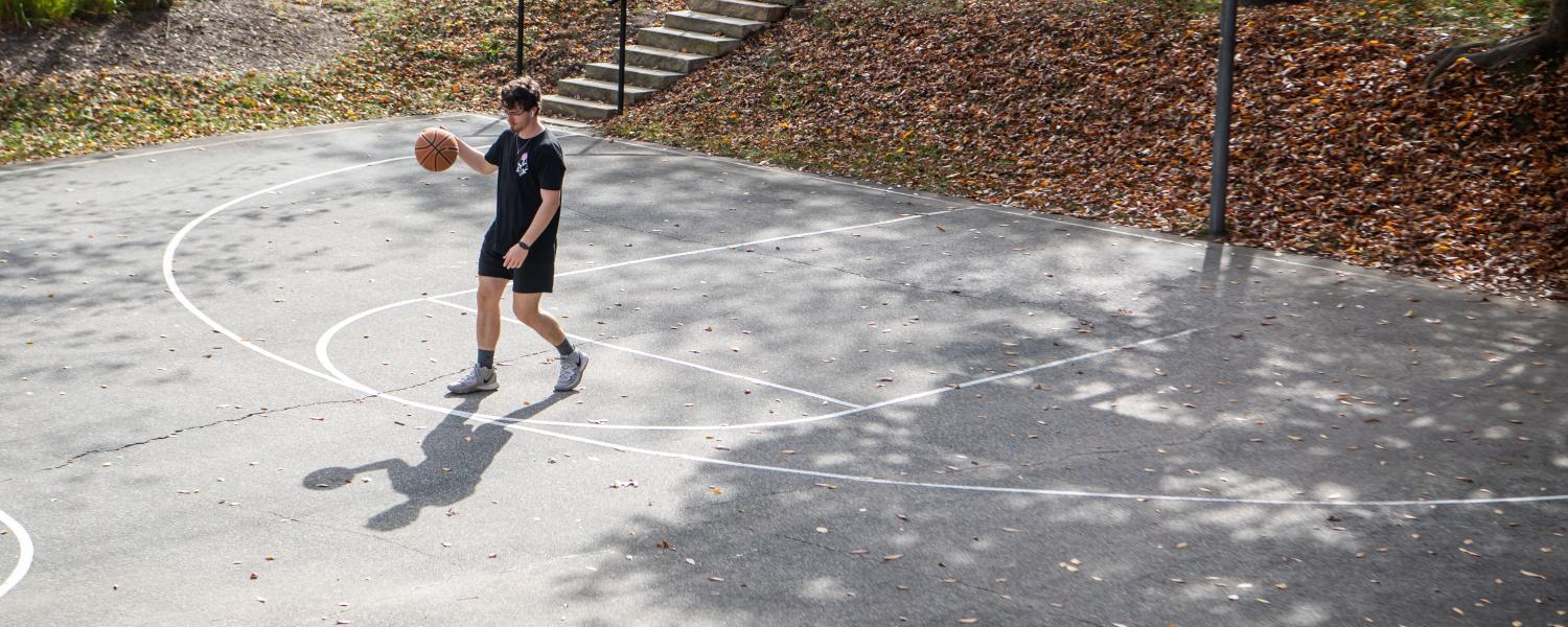 A student plays basketball alone on an outdoor court while listening to music through earbuds.