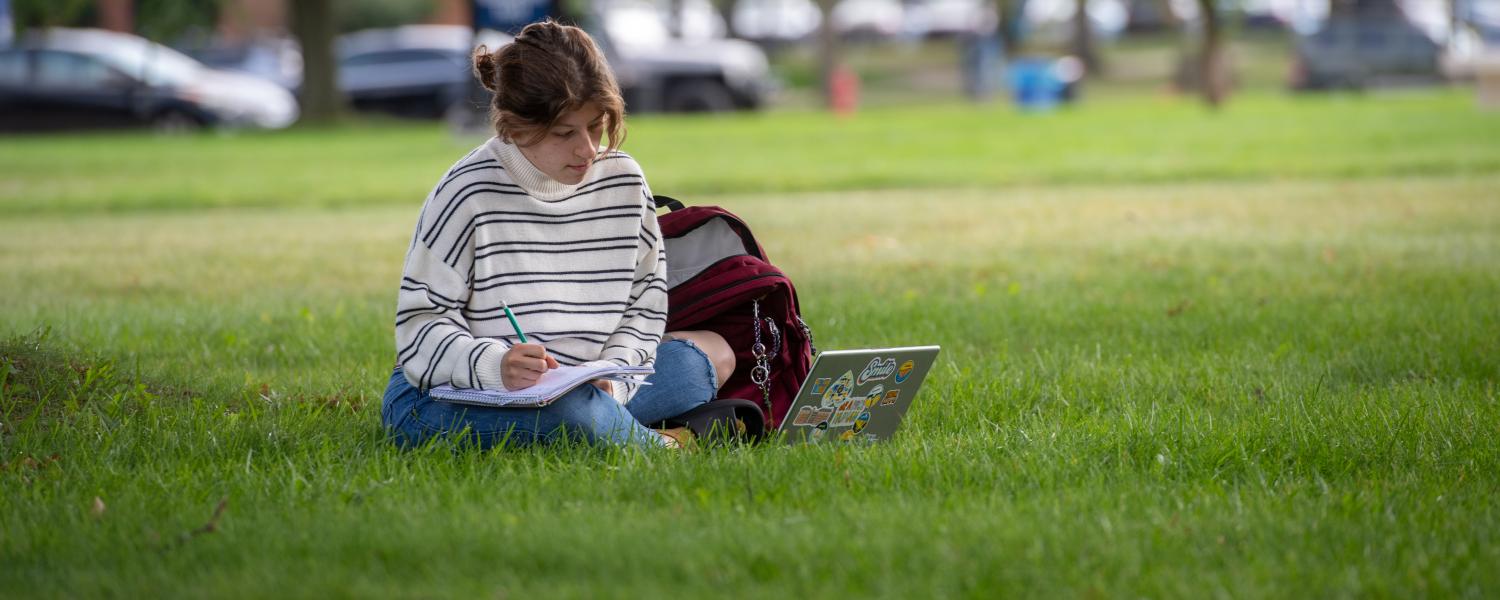 A student sits in the grass on a sunny day, studying with a notebook and a laptop