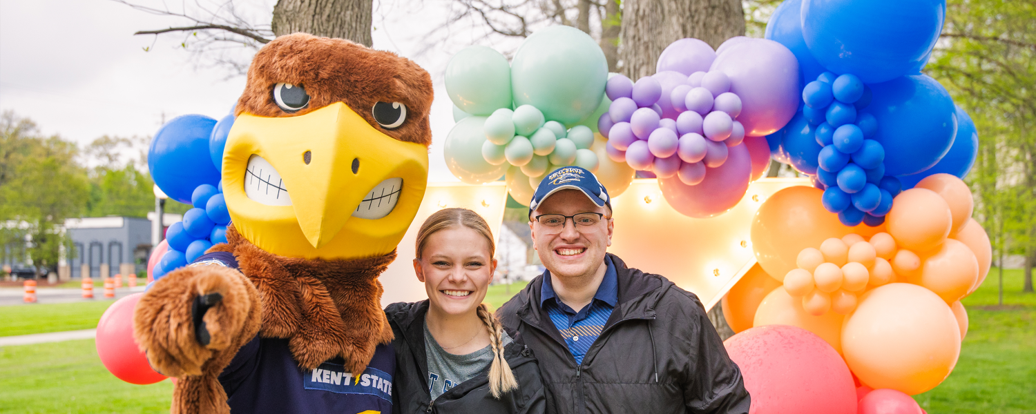 Flash poses with two Kent State students with a colorful balloon background behind them.
