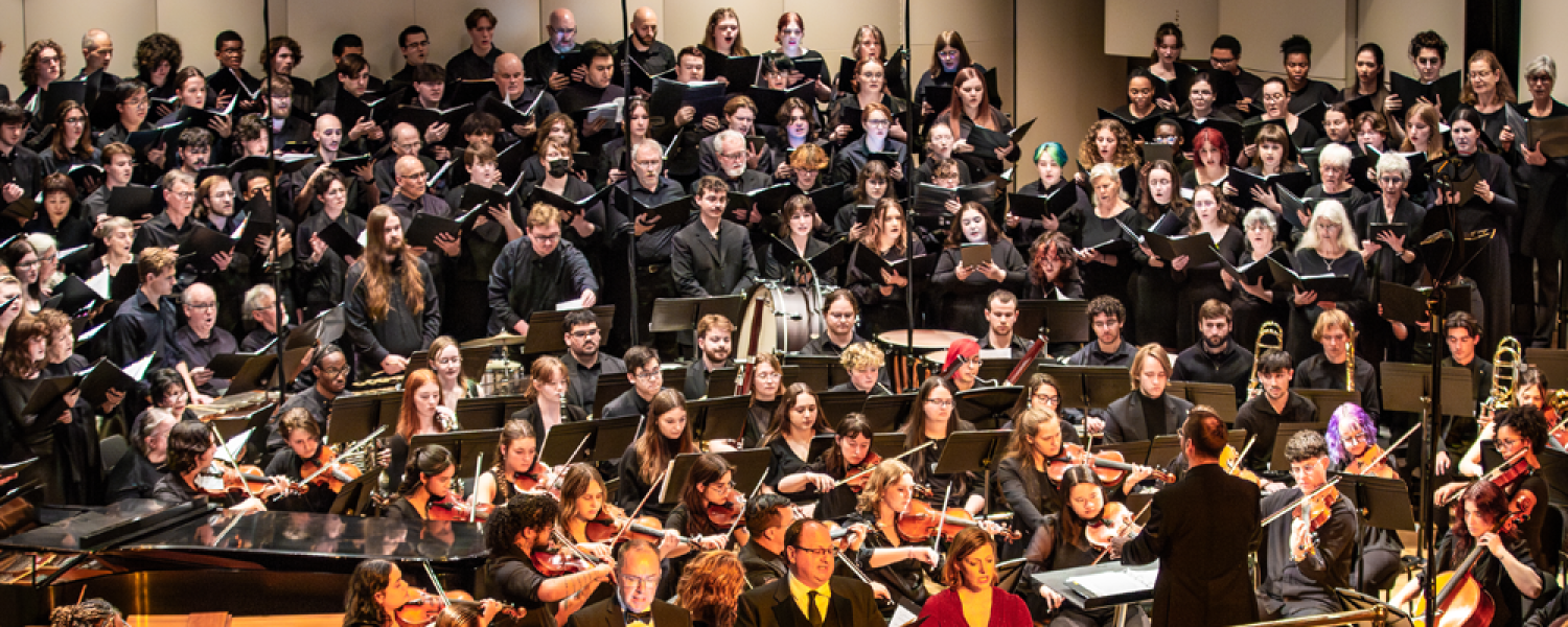 A large group of musicians prepare to play instruments on stage in front of a large group of choral vocalists in back