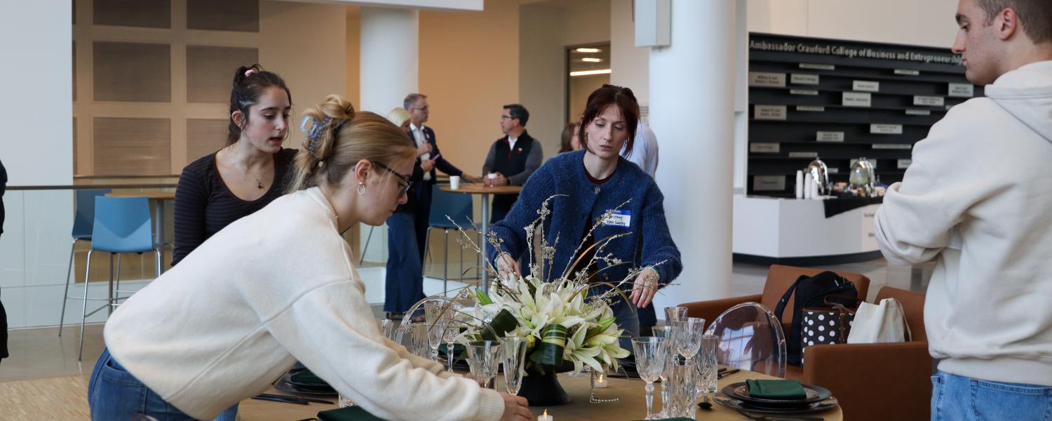 Teams of students and club managers adjust place settings on a table during a design challenge.