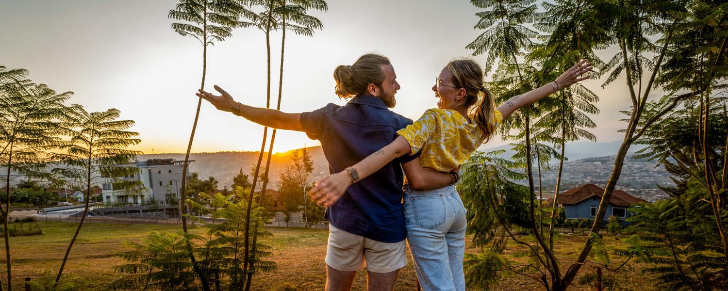 A golden-hour medium shot of a man and a woman standing in a grassy area with their backs partially to the camera, embracing and looking at each other joyfully with arms wide open. The setting sun glows behind them through thin, tall trees, overlooking a valley with houses scattered across the hills in the background.