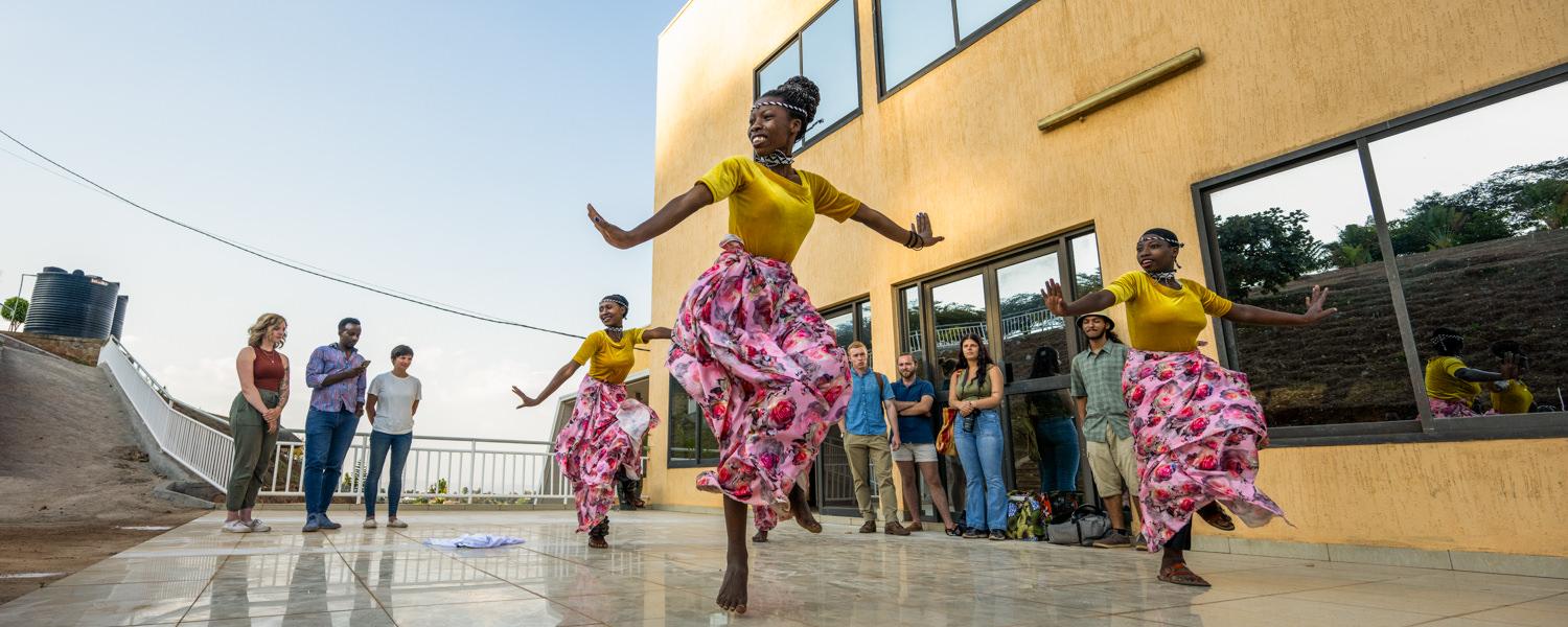 A wide shot of three Rwandan women performing a traditional dance on a polished, reflective outdoor patio next to a modern yellow building. They are wearing bright yellow tops and vibrant pink-and-white floral skirts, captured mid-movement with arms outstretched. In the background, a group of students and faculty members stand watching the performance.