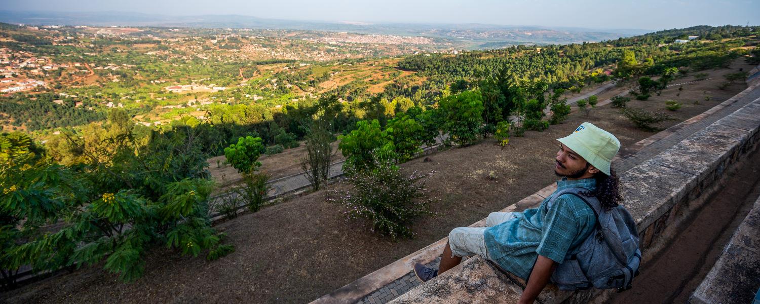 A high-angle, wide shot of a young man with curly dark hair wearing a light green bucket hat, a blue plaid button-down shirt, and a backpack. He is sitting on a stone ledge, looking out over a massive, lush green valley that stretches toward a distant city under a soft haze.