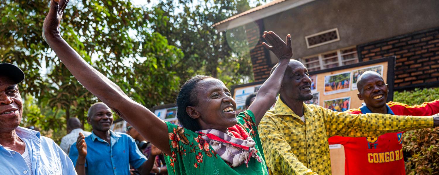A vibrant, close-up shot capturing a moment of celebration. A Rwandan woman in a green floral dress has both arms raised high in the air, smiling brightly. Surrounding her are several other men and women laughing and cheering, with a wooden display board featuring photographs visible in the background.