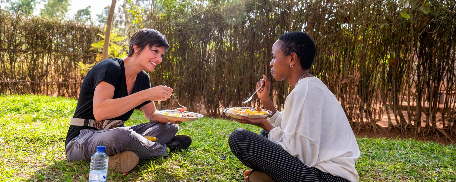 A medium shot of two women sitting cross-legged on a lush green lawn, enjoying a meal together outdoors. On the left, a Caucasian woman with short dark hair in a black T-shirt smiles warmly at a Rwandan woman with short hair on the right, who wears a white blouse and striped trousers. Both hold plates of food and forks, with a rustic woven branch fence behind them.