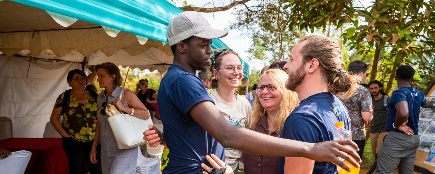A joyful group interaction outdoors under a bright blue-and-white canopy. In the foreground, a young Rwandan man wearing a baseball cap and a dark blue shirt is embracing or greeting a Caucasian man with long hair tied back, who is also wearing a С��Ƭ��Ƶ blue shirt. Surrounding them are other smiling students and faculty members.
