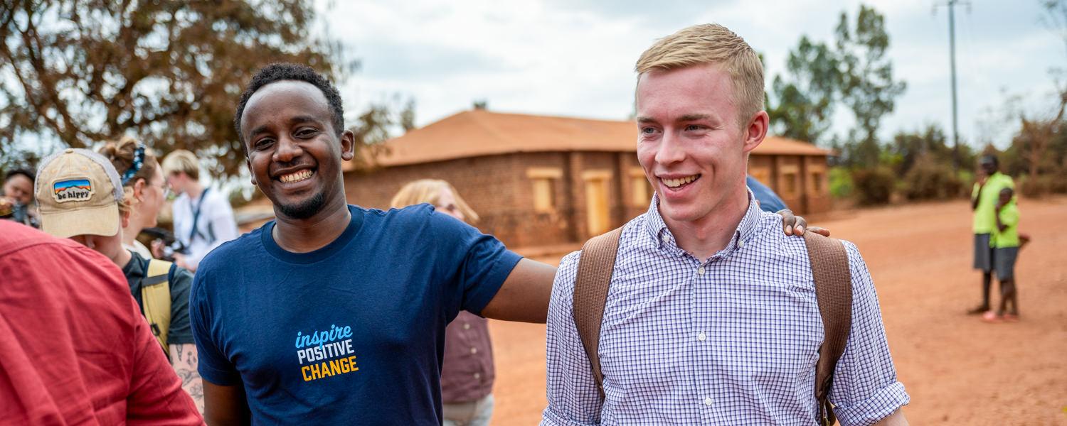 candid medium shot of two young men smiling and walking together on a dirt road. On the left, a Rwandan man in a dark blue T-shirt that reads "inspire POSITIVE CHANGE" smiles broadly with his arm around a blond Caucasian man wearing a purple gingham button-down shirt and a backpack. Other people and a simple brick building are visible in the background.